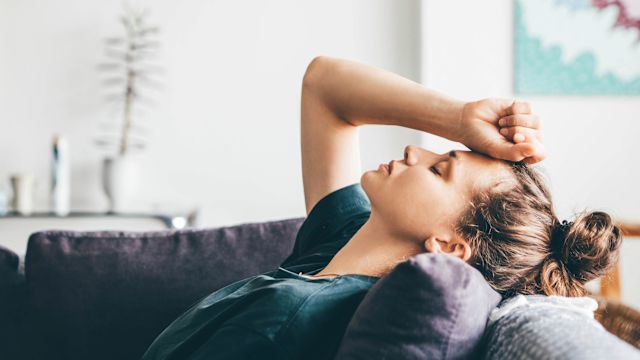 An exhausted young woman rests on a couch with her hand on her head. Fatigue is a common symptom of PNH and can have multiple contributing factors.