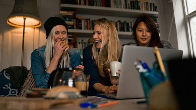 Three young women are sitting on a couch in an apartment. They are watching something on a computer and laughing.