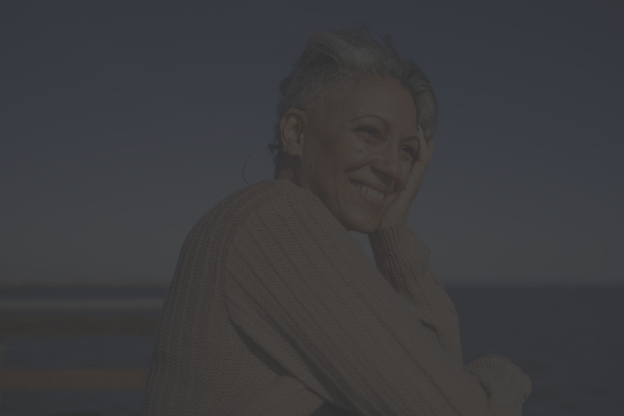 a middle aged Black woman smiles and looks off to sea on a sunny day near the beach