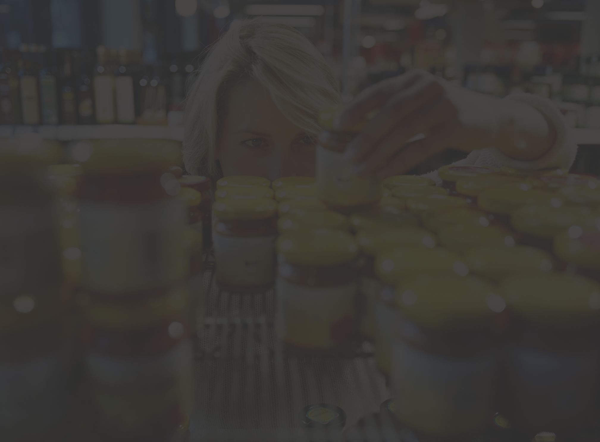 a white woman with blonde hair peers at a shelf of jarred condiments, reading the nutrition labels to find out how much added sugar they contain