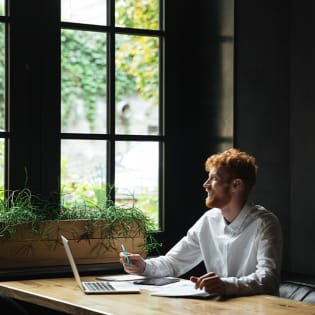 A smiling red-haired man in a white shirt sits at a wooden table with a laptop near a large window.