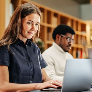 A woman in a dark shirt types on a laptop in a library with a man working in the background.